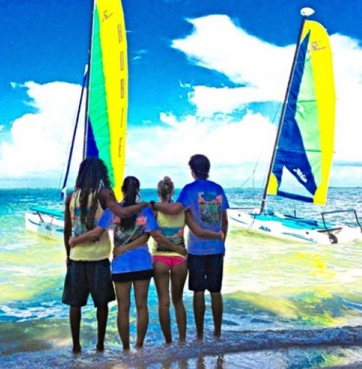 Four kids standing arm in arm on the shore watching Hobie catamarans sail past