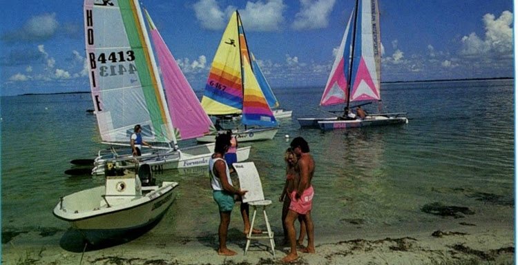 An artist painting at an easel on the beach as Hobie catamarans come ashore behind her