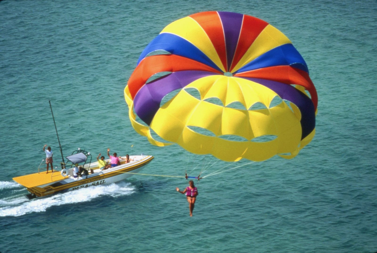 Aerial view of a rainbow parasail flying behind a yellow towboat over green water